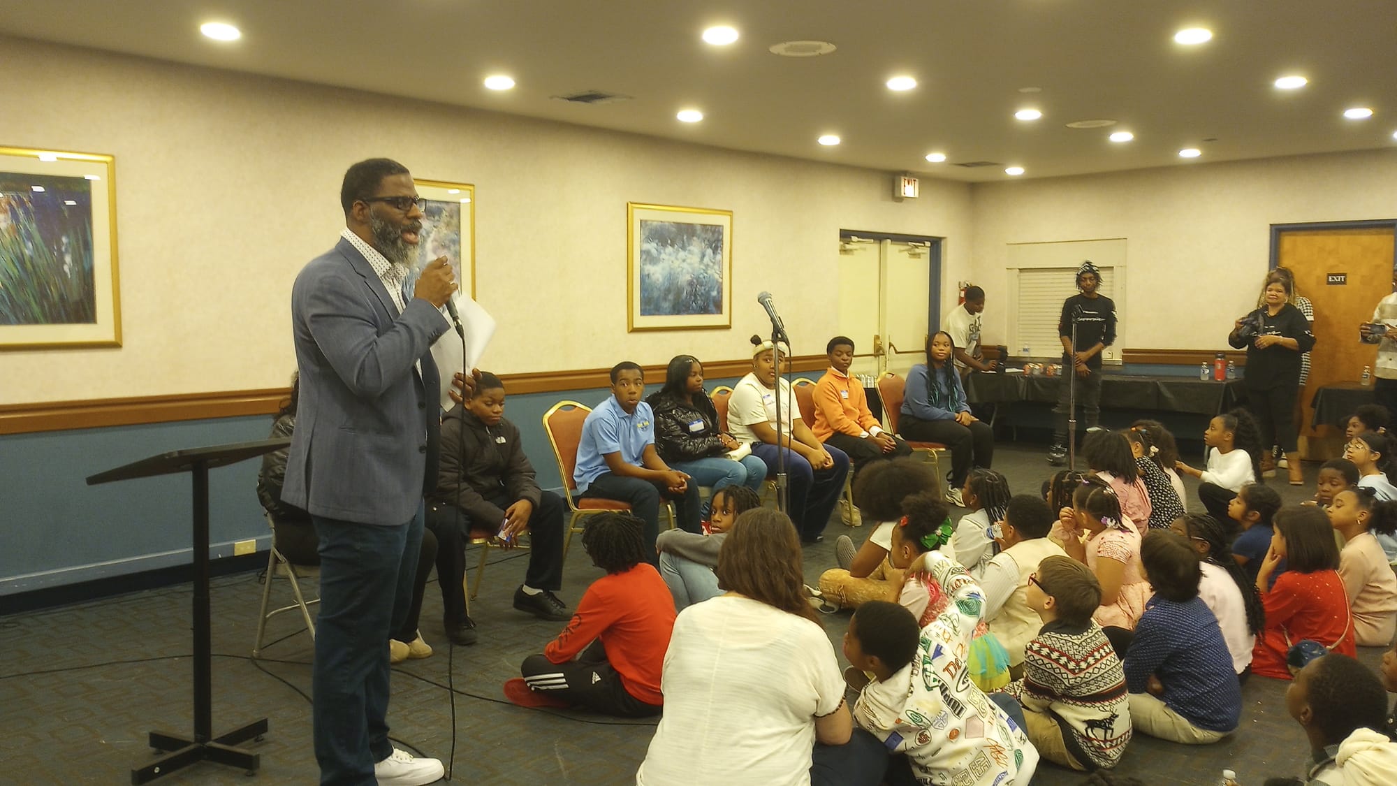 The image shows board member Che "Rhymefest" Smith standing, holding a microphone, next to a panel of six students. Chicago Board of Education Honorary Student Member Destiny Singleton is seated at the far right of the panel.