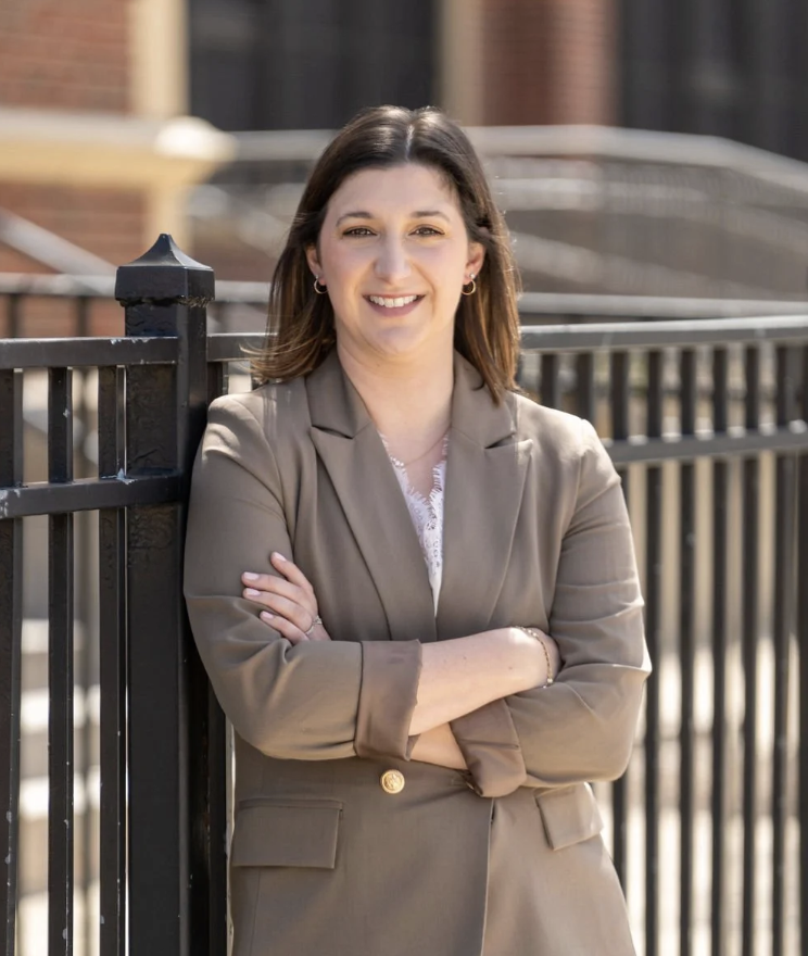 The image shows Chicago Board of Education member Jennifer Custer leaning against an iron fence. Her arms are crossed. A school building is in the background.