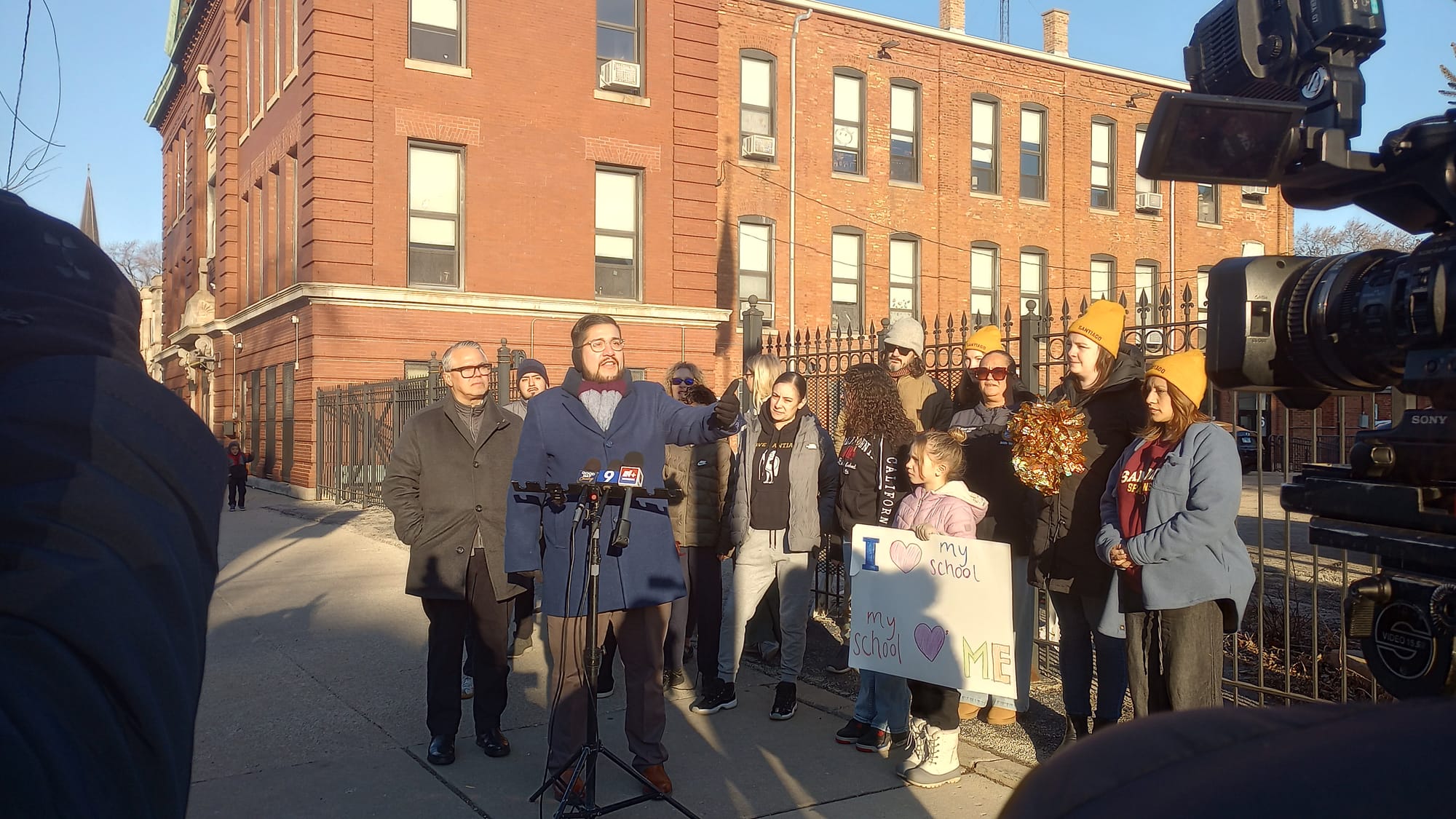 Carlos Rivas, in a blue coat, stands in front of a stand of multiple microphones. Ald. Gil Villegas stands behind him (left) and parents and students stand to the right.