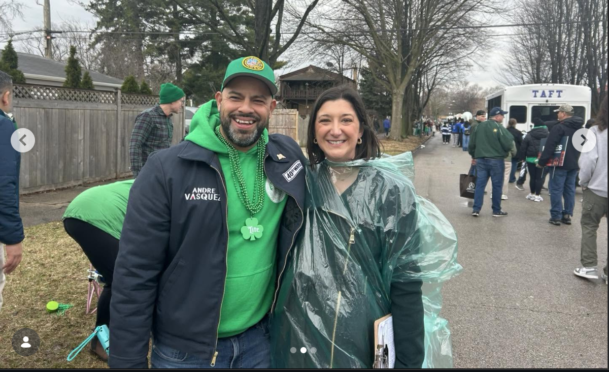 From left: Alderman Andre Vazquez (bearded, wearing a green hat, green sweatshirt and green bead necklaces) stands next to school board Member Jennifer Custer (wearing a clear green plastic rain poncho over a dark green coat.)