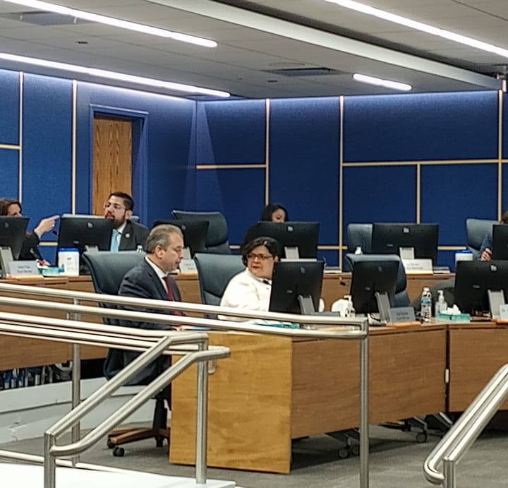 Chicago Board of Education members and staff chat, seated behind computer screens.