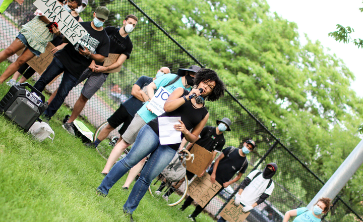 The photo centers on Cydney Wallace holding a microphone. Protestors holding Black Lives Matter signs stand behind her.