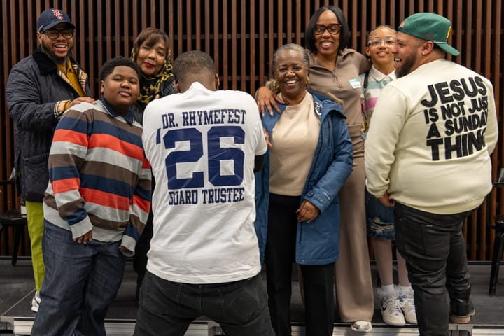 The photo shows a group of parents and young people, with Che "Rhymefest" Smith in the center-left. His back is turned to the camera and he wears a t-shirt: Dr. Rhymefest '26 Board Trustee.