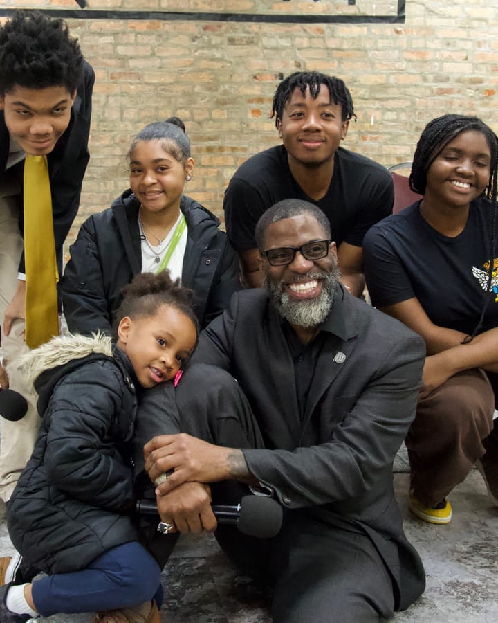 The photo shows board member Che "Rhymefest" Smith (center) holding a microphone and surrounded by young people.